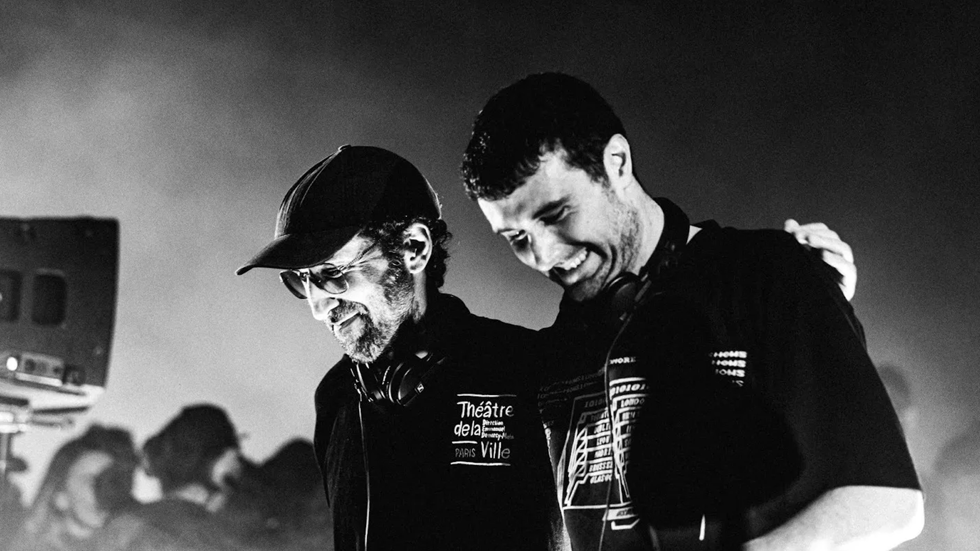 A black and white photo shows two smiling men, both with headphones around their necks; one bearded man in a cap and glasses, with the other's arm around his shoulder.