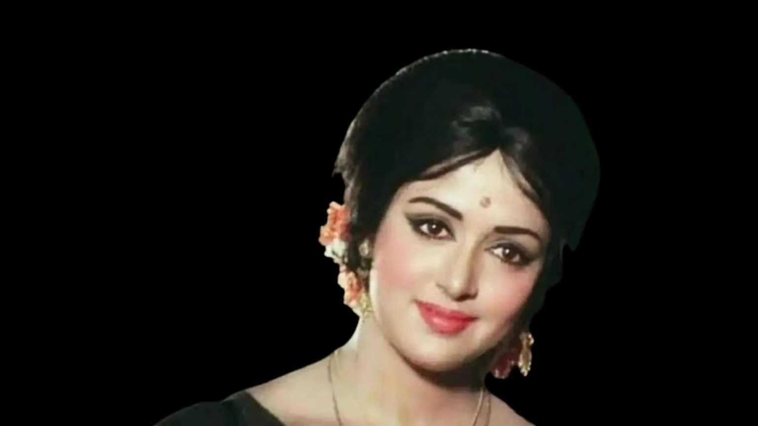 Close-up of a smiling woman with dark hair styled with orange flowers, winged eyeliner, a bindi, and a gold necklace, against a black background.
