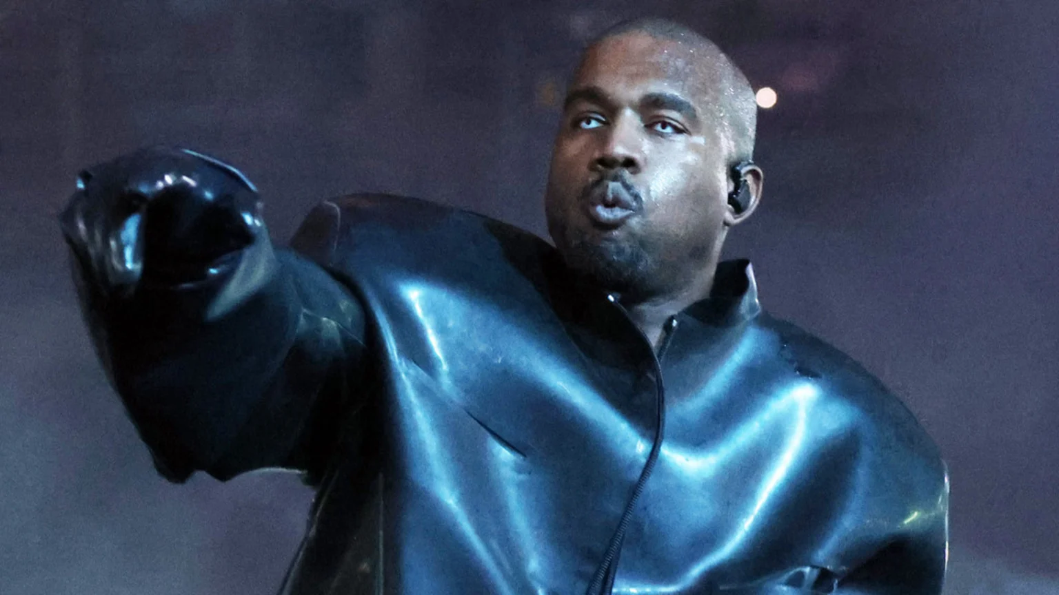 Kanye West, bald with a beard, wears a shiny dark jacket and has striking white eyes, with his lips pursed and an earbud in his ear, in a dim setting.