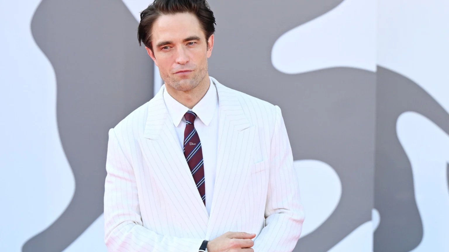 Robert Pattinson in a white pinstripe suit, white shirt, and a burgundy and navy striped Dior tie, looking at the camera against an abstract gray and white background.