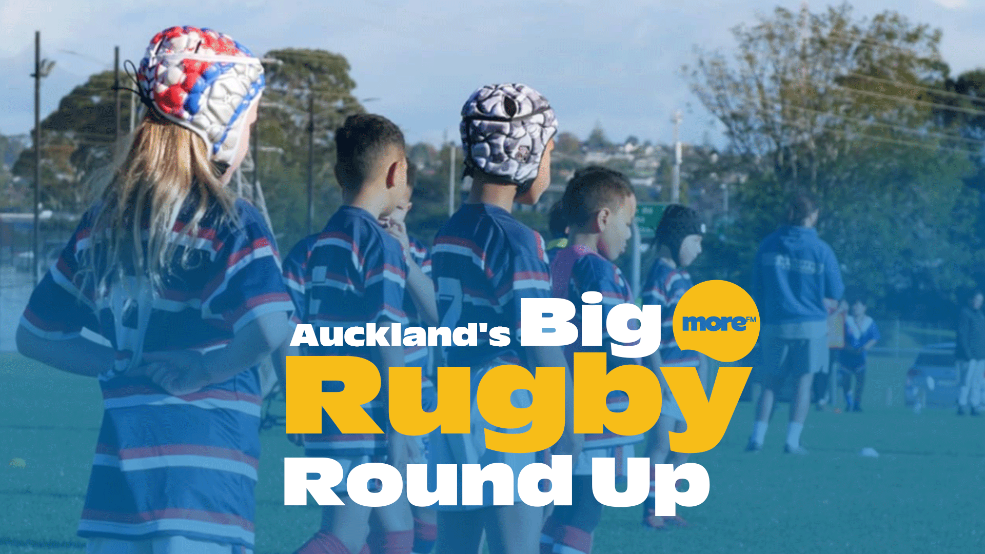 Children in rugby jerseys and headguards stand on a green field under a clear sky, with text "Auckland's Big Rugby Round Up" and "more FM" overlayed.
