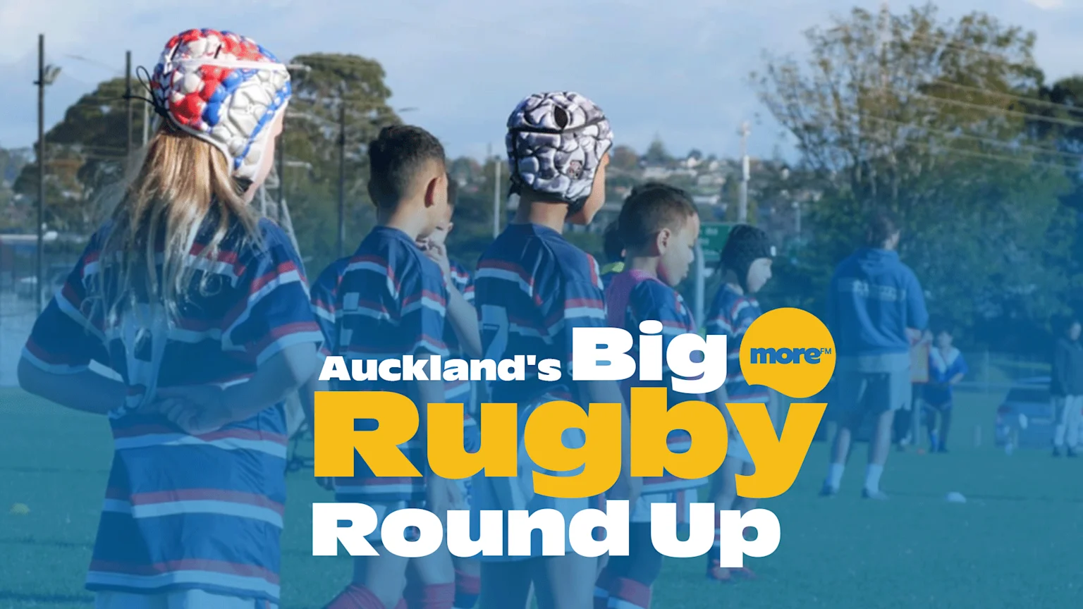 Children in rugby jerseys and headguards stand on a green field under a clear sky, with text "Auckland's Big Rugby Round Up" and "more FM" overlayed.
