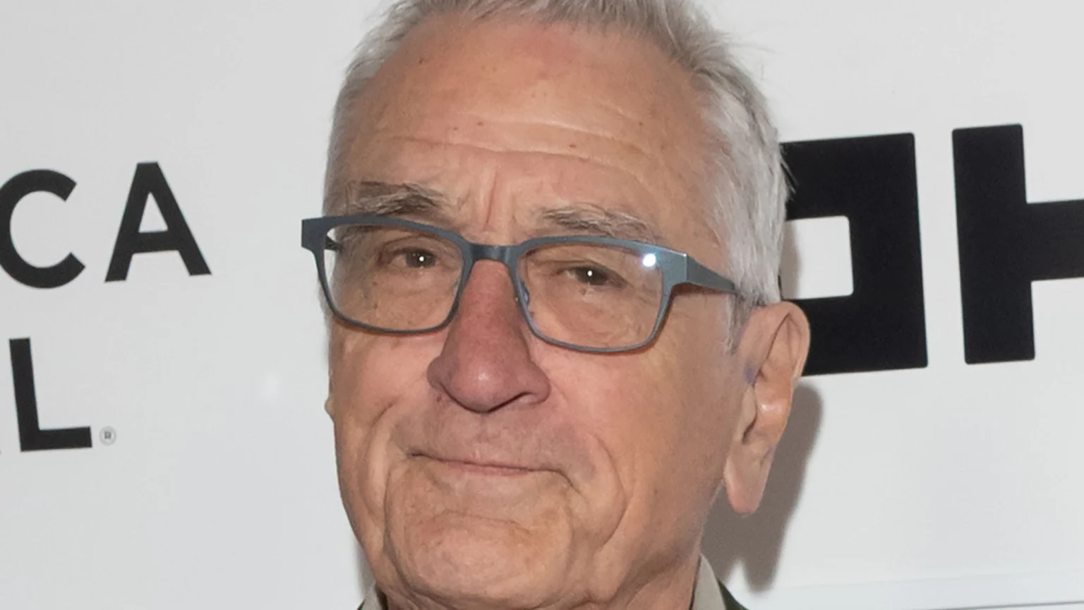 Close-up of Robert De Niro, an older man with grey hair and glasses, looking forward with a neutral expression against a white background with partial black text.