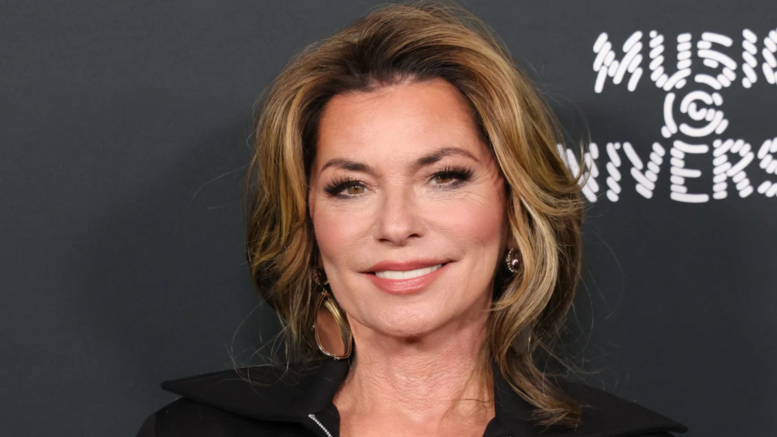 Shania Twain smiles in a close-up headshot, her blonde hair styled around her face, wearing noticeable makeup including eyelashes, earrings, and a dark top, against a grey background with white text.