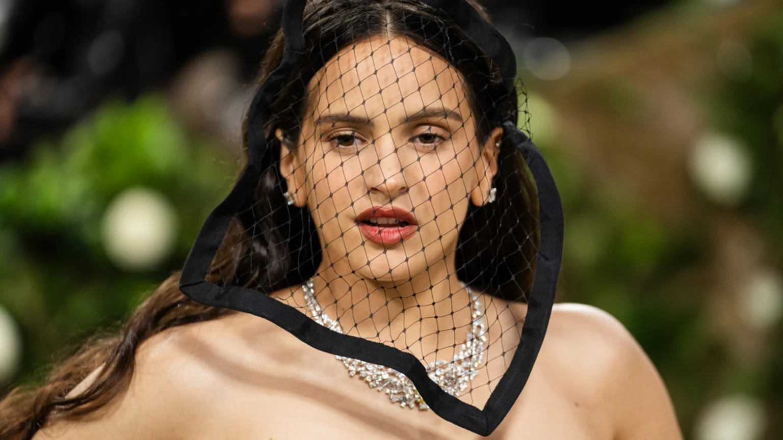 Close-up of Rosalía wearing a black mesh veil over her face, a diamond necklace, and stud earrings, against a blurred green background.