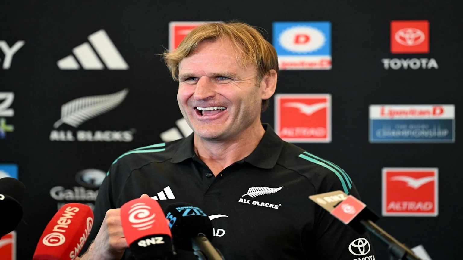 Scott Robertson, a fair-haired man, smiles while speaking at a press conference. He wears an All Blacks polo shirt, surrounded by microphones and sponsor logos on a dark backdrop.