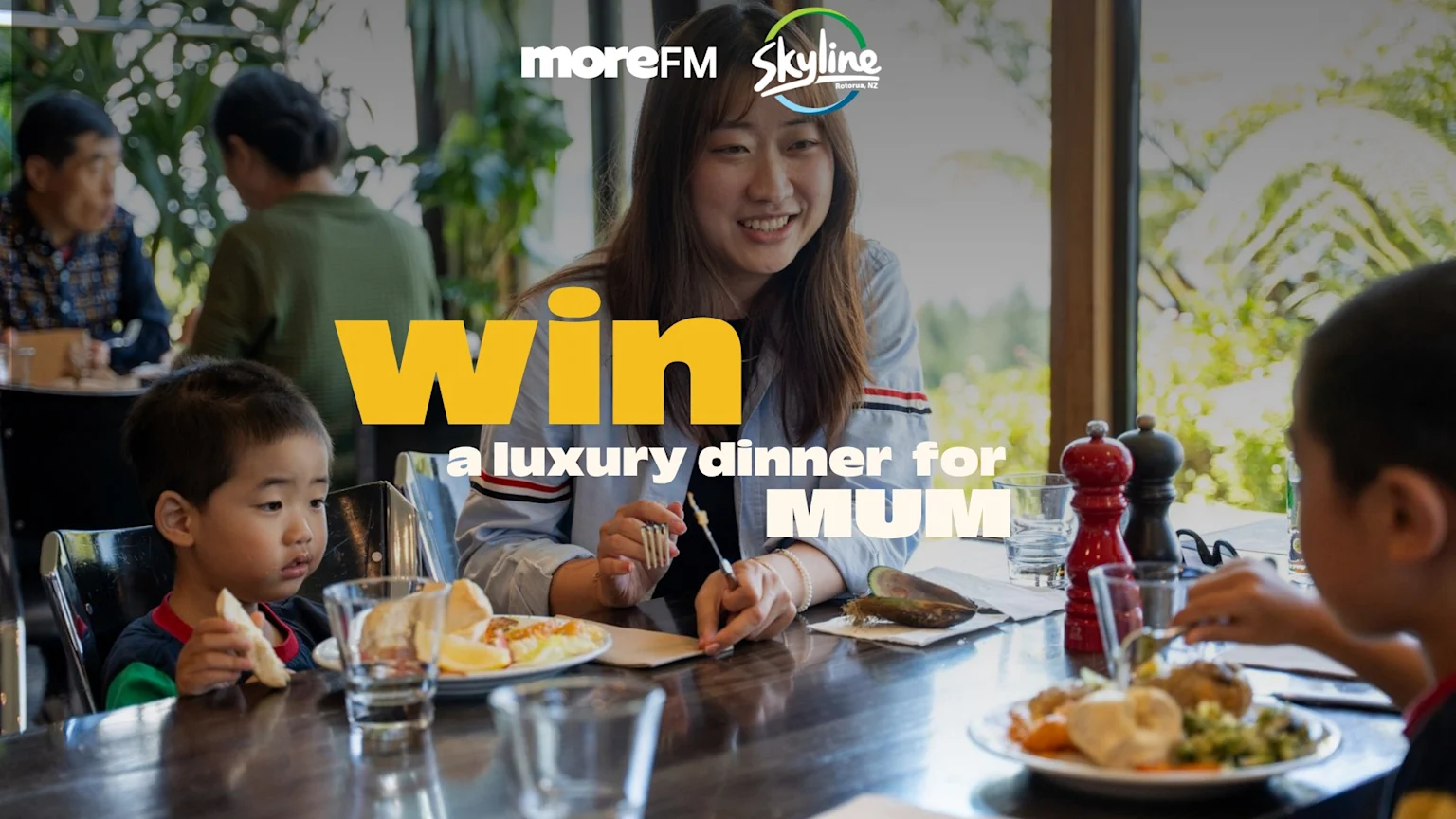 A smiling woman and two young children enjoy dinner at a restaurant, with text 'WIN a luxury dinner for MUM' and 'moreFM Skyline Rotorua, NZ' logos.