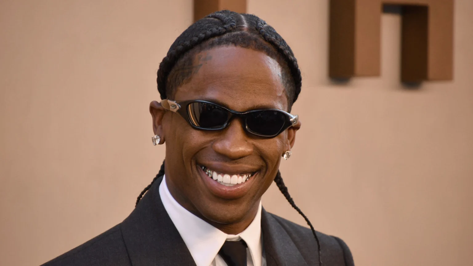 Travis Scott smiles widely, wearing black sunglasses, diamond stud earrings, a dark suit with a white shirt and black tie, and a grill on his bottom teeth, with braided hair.