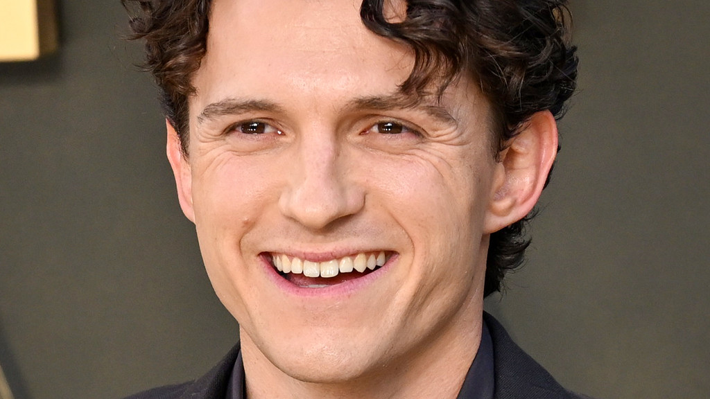Close-up of a man with dark, curly hair and brown eyes, smiling broadly to reveal his teeth, with crow's feet visible around his eyes.