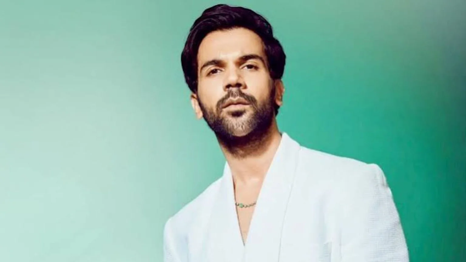 A man with dark hair, beard, and mustache, wearing a white blazer and a subtle green-beaded necklace, looks intently at the viewer against a gradient mint green background.