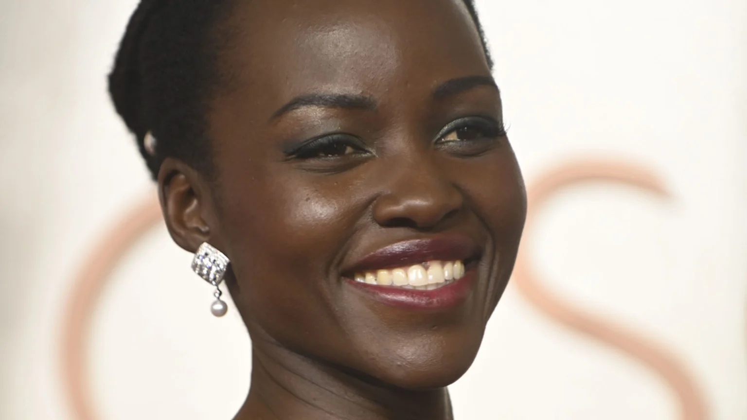 Lupita Nyong'o smiles broadly, her dark skin glowing, wearing dark red lipstick and a sparkling diamond and pearl earring on her left ear.
