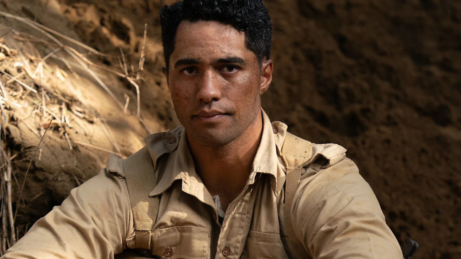 A Māori soldier with dark curly hair and a dusty face wears a tan military uniform, looking intently at the viewer in a dirt trench.