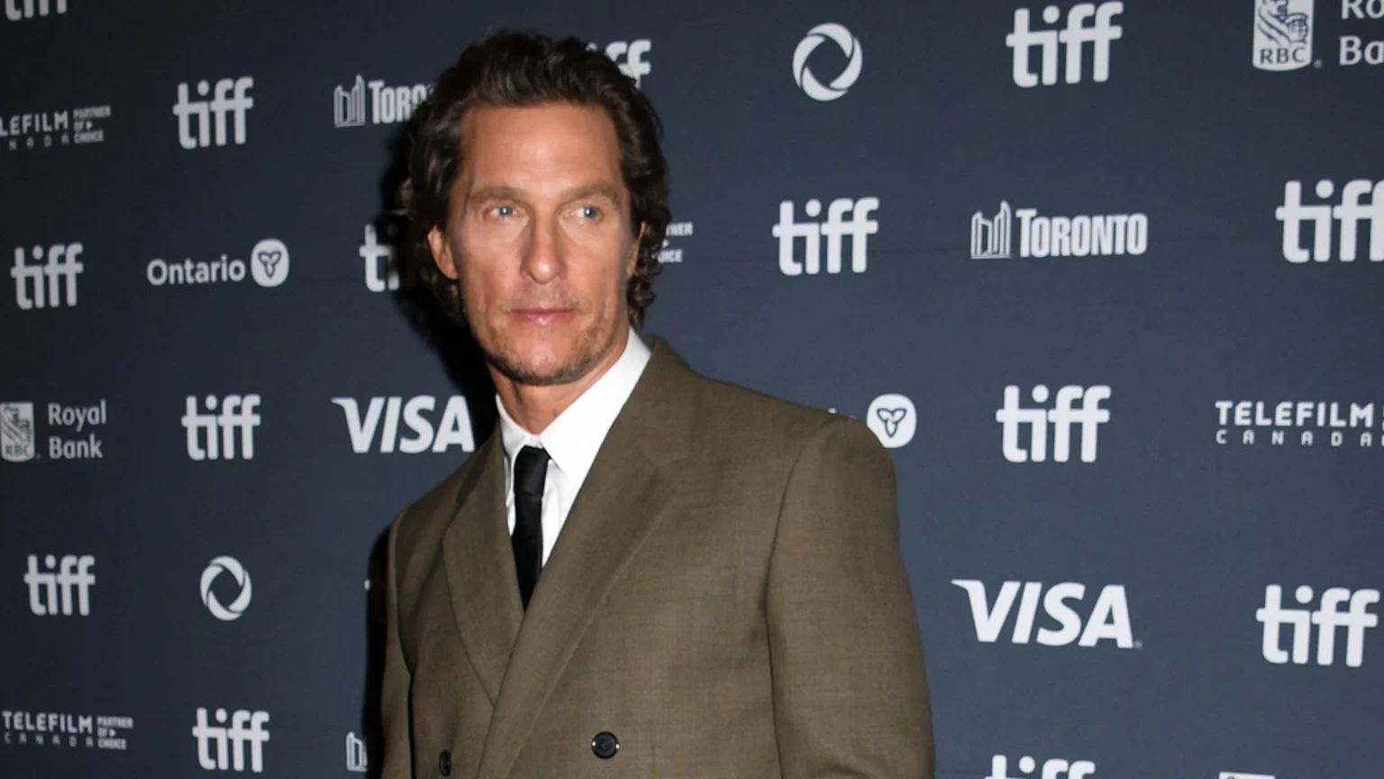 Matthew McConaughey in a brown suit, white shirt, and black tie, stands against a dark background with white "TIFF" and other logos, looking slightly left.