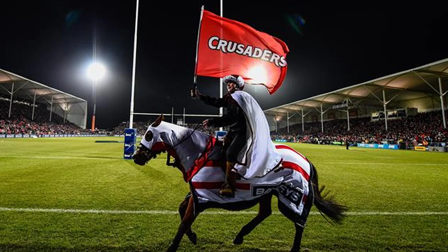 A person in a white cape rides a horse adorned in white and red, carrying a "CRUSADERS" flag, across a brightly lit rugby stadium at night with spectators in the stands.