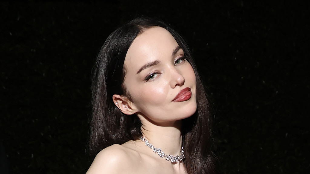 Dove Cameron, with long dark hair, dark red lipstick, a sparkly choker necklace, and an ear piercing, poses looking to her right with a subtle expression, her bare shoulder visible against a dark background.