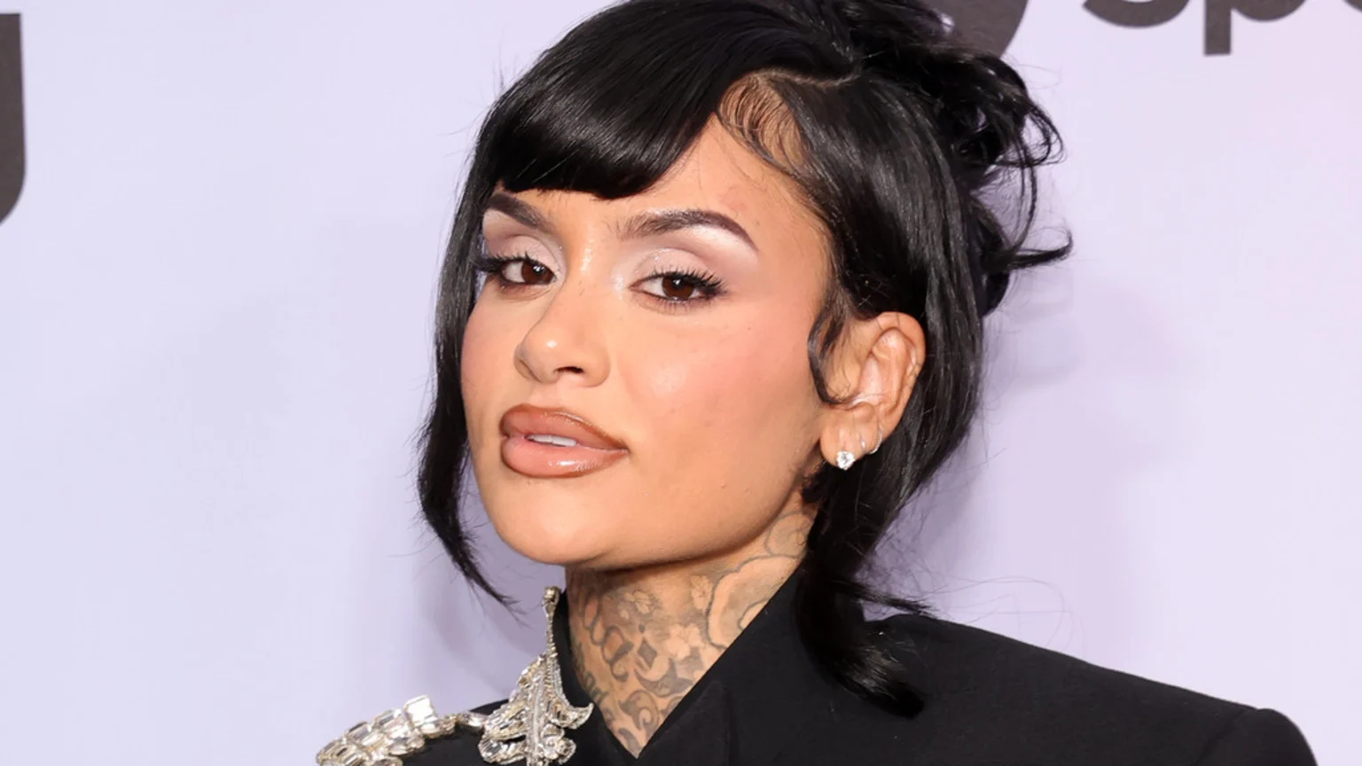 Close-up portrait of Kehlani with dark hair, bangs, makeup, and neck tattoos, wearing a black top with silver embellishments.