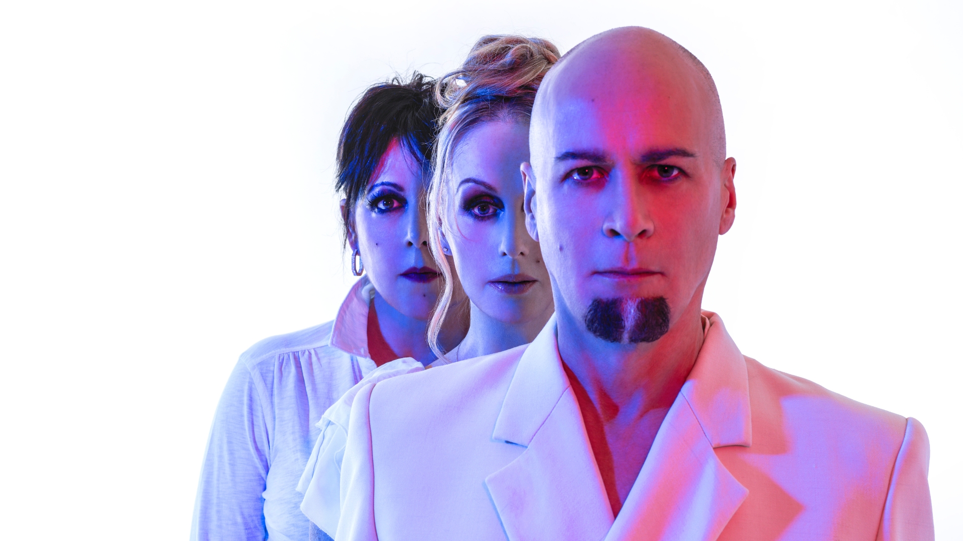 Three people—a man, a blonde woman, and a dark-haired woman—stand in a line, looking directly at the viewer, bathed in red and blue lights against a white background.
