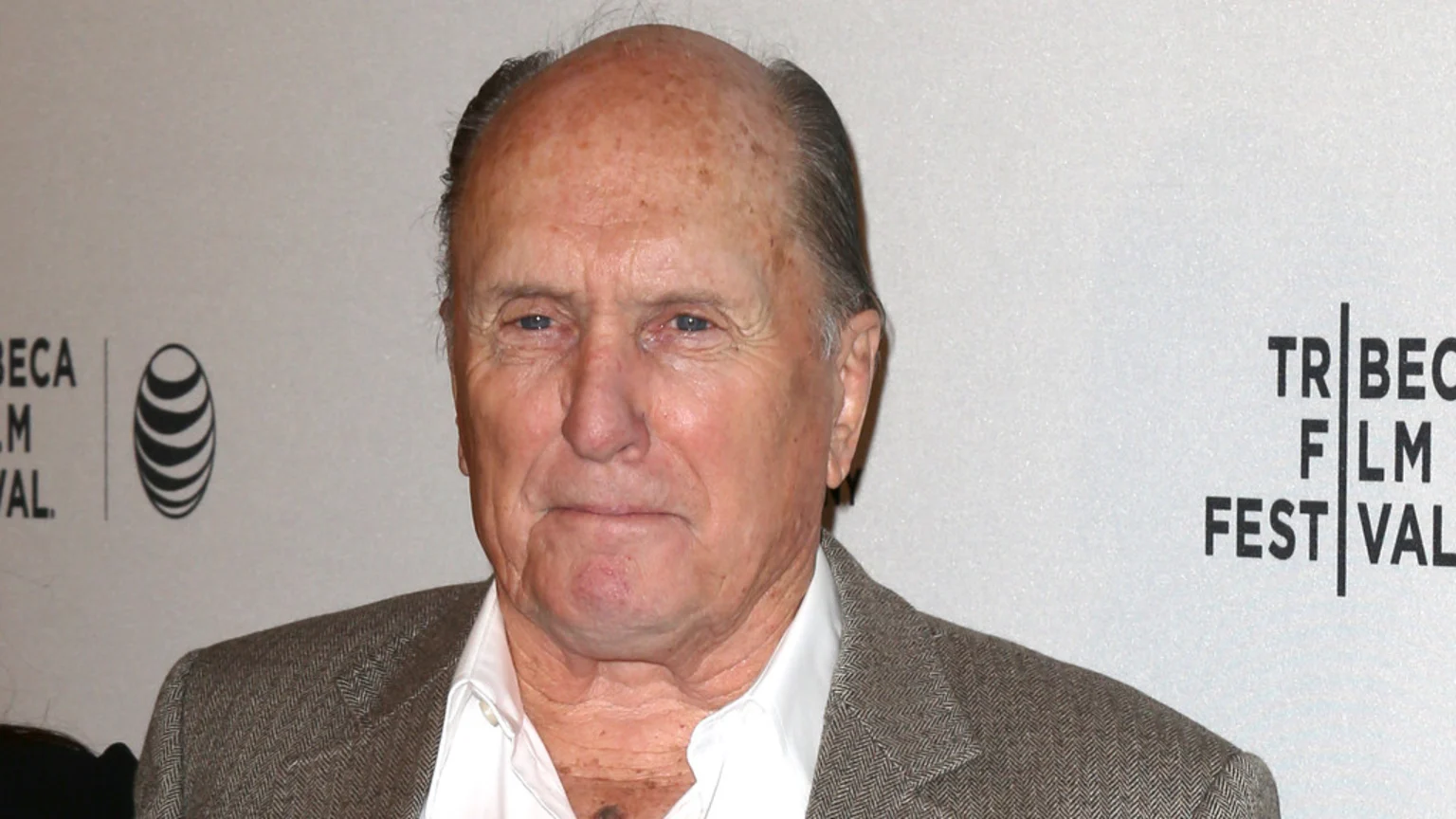 Close-up portrait of Robert Duvall at the Tribeca Film Festival, wearing a white collared shirt and brown blazer, standing indoors with bright lighting in front of branded walls featuring the AT&T black-and-white globe logo.