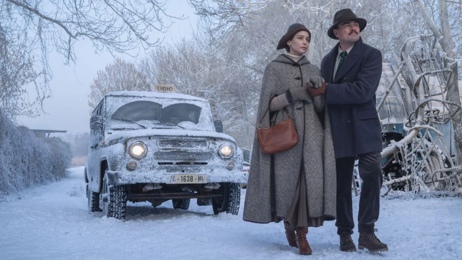 A man resembling Leonardo DiCaprio and a woman in winter attire, holding hands, stand on a snowy path with a snow-covered utility vehicle and frosty trees in the background.