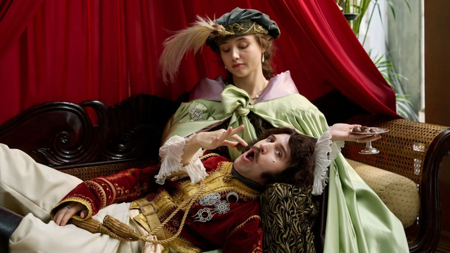 A woman in a green gown and feathered hat offers a chocolate to a man in a red and gold jacket who is lying on her lap on a sofa, with red curtains behind them.