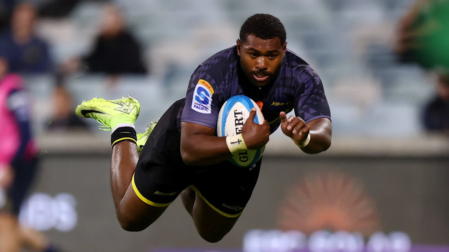 A male rugby player with dark skin and hair, in a patterned dark jersey with a DHL Super Rugby Pacific logo, black shorts, and bright yellow cleats, dives through the air, clutching a blue and white Gilbert rugby ball, with a blurred stadium crowd behind.