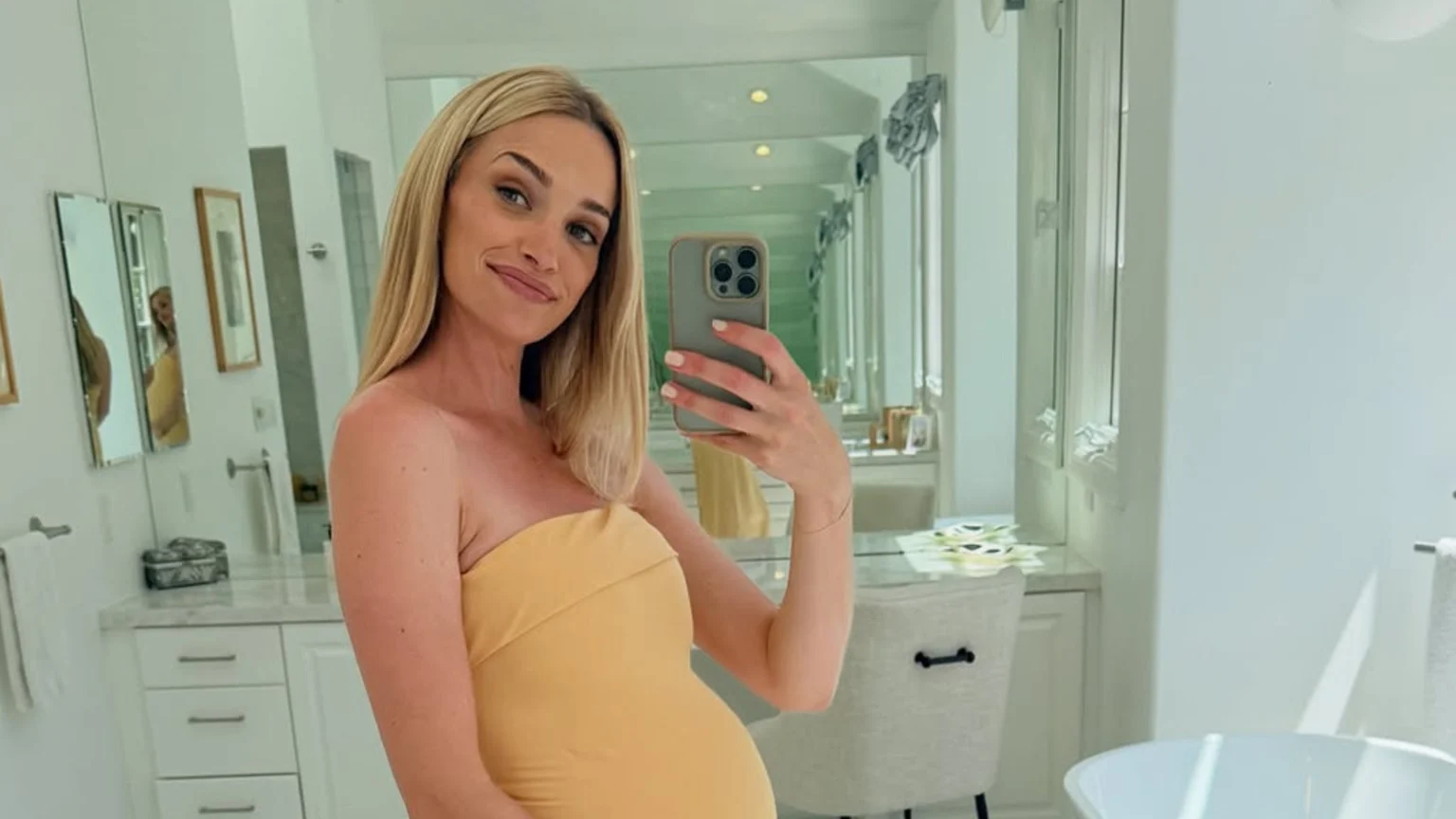 A pregnant woman with blonde hair in a yellow strapless dress takes a mirror selfie, smiling and holding her belly in a bright bathroom.