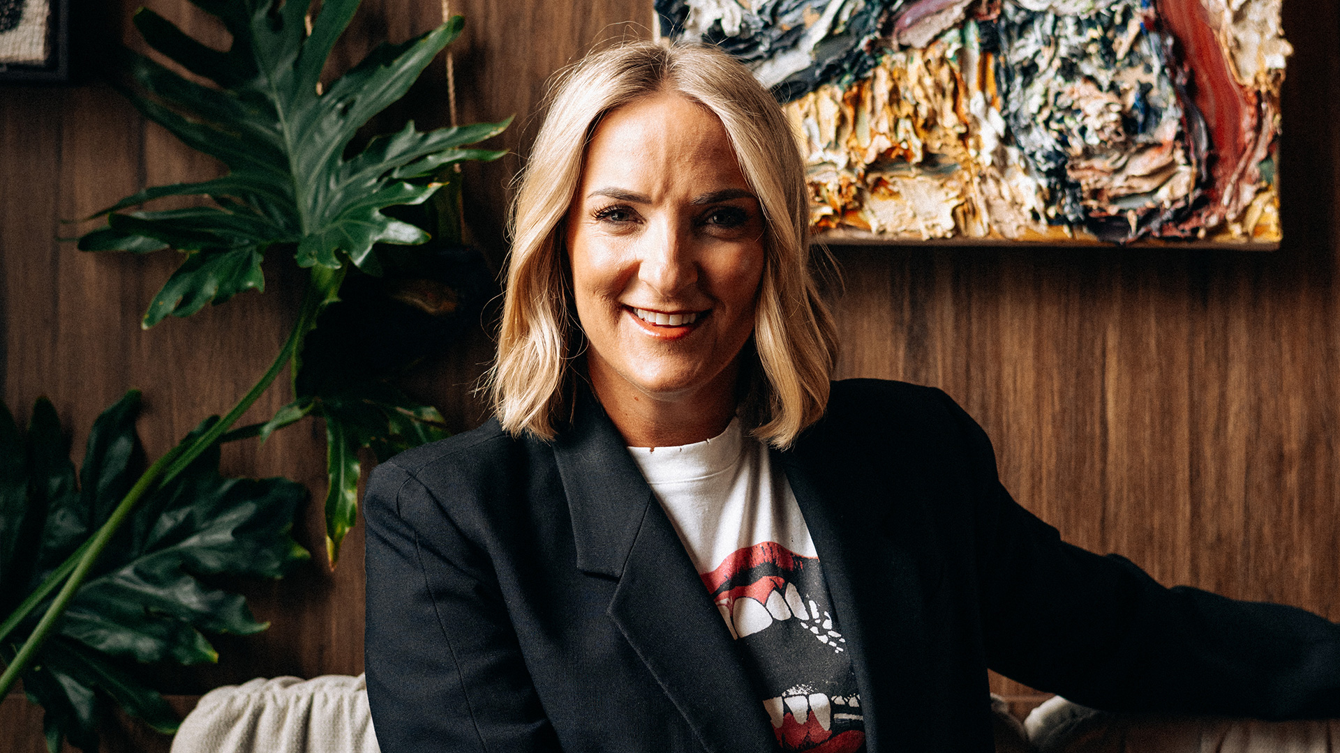 A smiling blonde woman with shoulder-length hair wears a black blazer and graphic t-shirt, sitting against a wooden wall with plants and abstract art in the background.