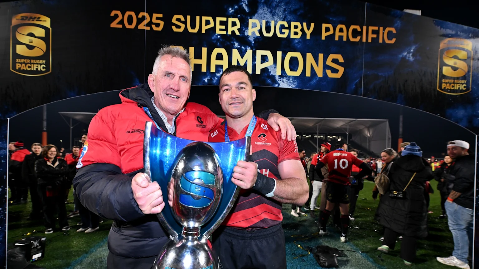 A Crusaders player and another man hold a silver and blue Super Rugby Pacific trophy, smiling in front of a banner reading "2025 SUPER RUGBY PACIFIC CHAMPIONS".
