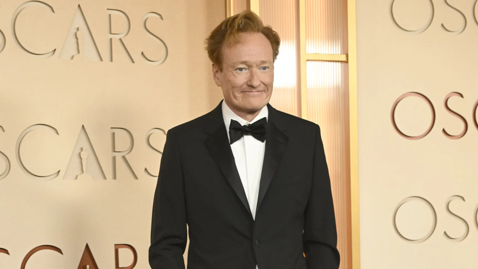 Conan O'Brien in a black tuxedo and bow tie, smiling in front of a light-colored wall with embossed "OSCARS" text and statuettes.