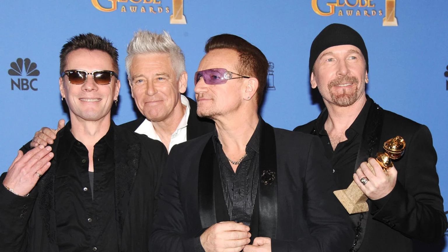 The rock band U2 poses together, with The Edge holding a Golden Globe award, against a blue backdrop featuring "Golden Globe Awards" and "NBC" logos.