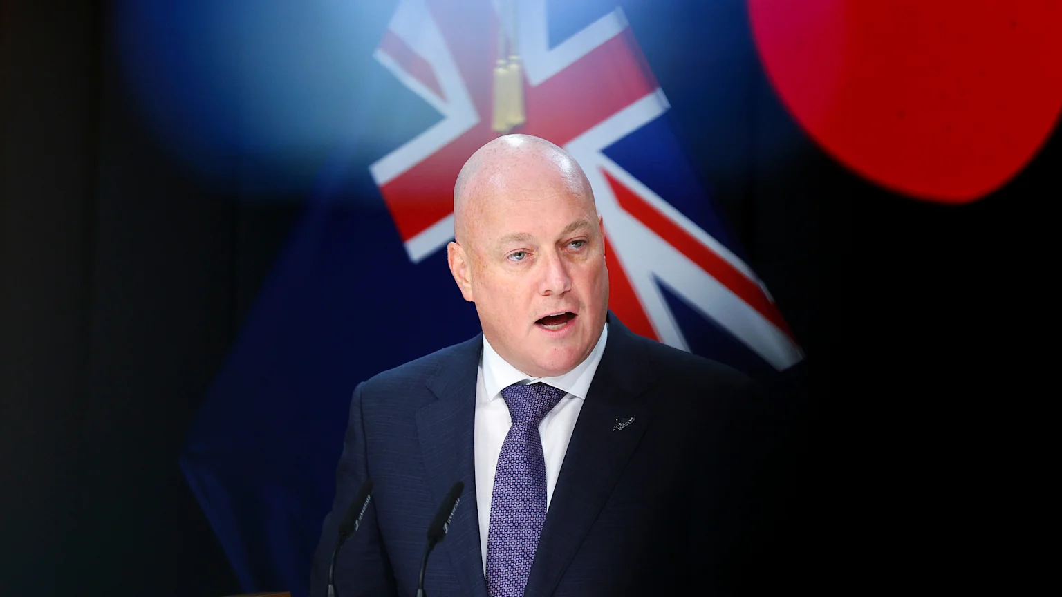 New Zealand politician Christopher Luxon, bald and wearing a dark suit and purple tie, speaks at a podium with microphones, with the blurred New Zealand flag and a red shape behind him.
