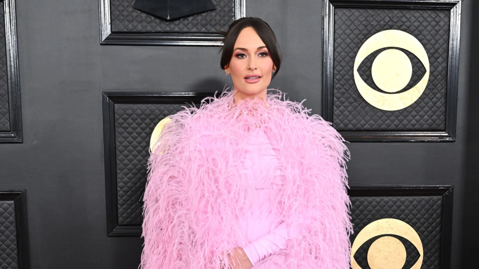 Kacey Musgraves in a fluffy pink outfit stands in front of a dark wall with framed CBS Eye logos.