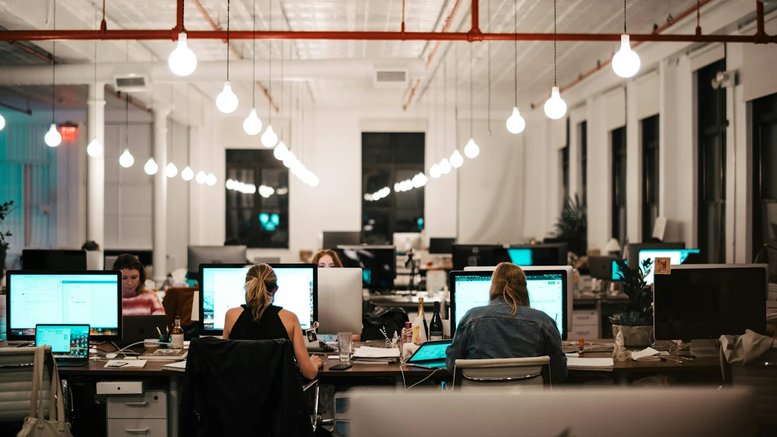 People work on individual computer workstations in a modern, brightly lit open-plan office, with rows of hanging light bulbs overhead.