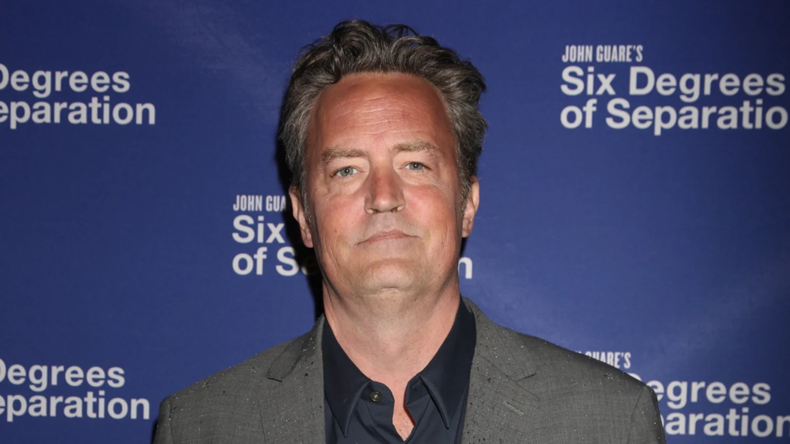 Matthew Perry, a middle-aged man with grey hair, wearing a grey suit and dark shirt, stands in front of a blue background with the white text "JOHN GUARE'S Six Degrees of Separation".