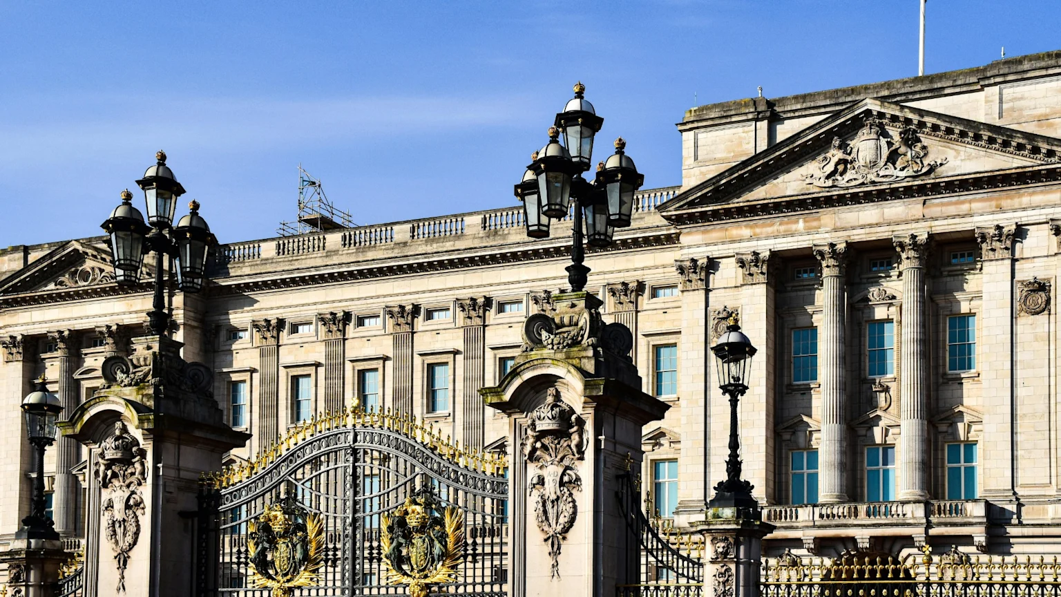 Buckingham Palace's grand stone facade is visible behind ornate black and gold gates and elaborate lampposts, set against a clear blue sky, with scaffolding on a distant part of the roof.