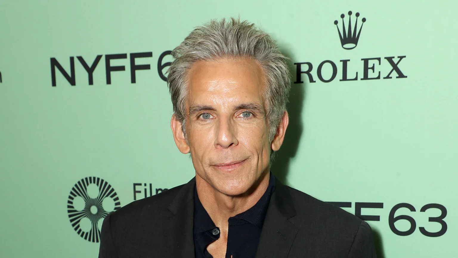 Ben Stiller, a man with gray hair and blue eyes, poses in a dark suit against a light green background with "NYFF" and "ROLEX" logos.