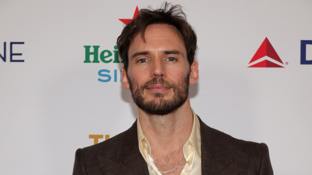 A man with a beard and brown hair wears a brown jacket over a light, open-collared shirt, featuring a silver chain, against a white wall with "Heineken" and "Delta" logos.