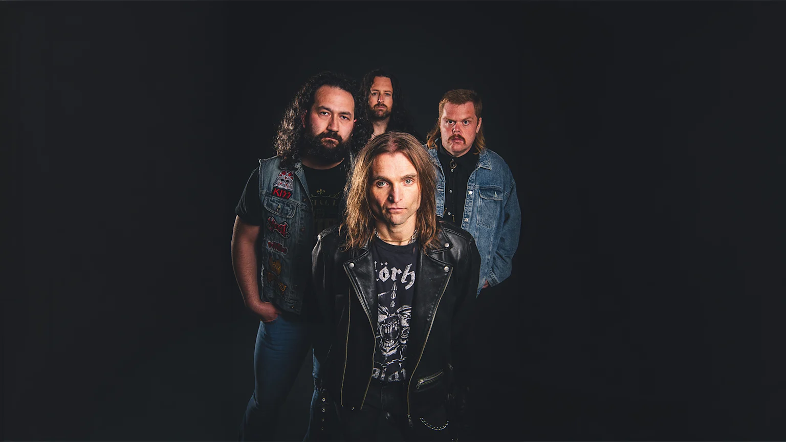 Four men with long hair, beards, and mullets, dressed in rock-style clothing including leather jackets and denim vests, pose seriously against a dark background.