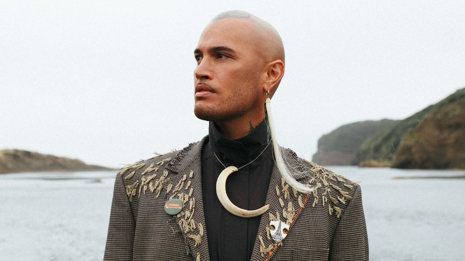 A bald man with a dark turtleneck and patterned jacket with fringe, wearing a large crescent necklace, looks left by water and hills under a grey sky.
