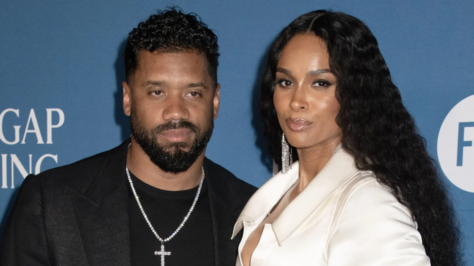 Singer Ciara, in a white top and long dark hair, stands next to Russell Wilson, in a black jacket and beard with a cross necklace, against a blue background.