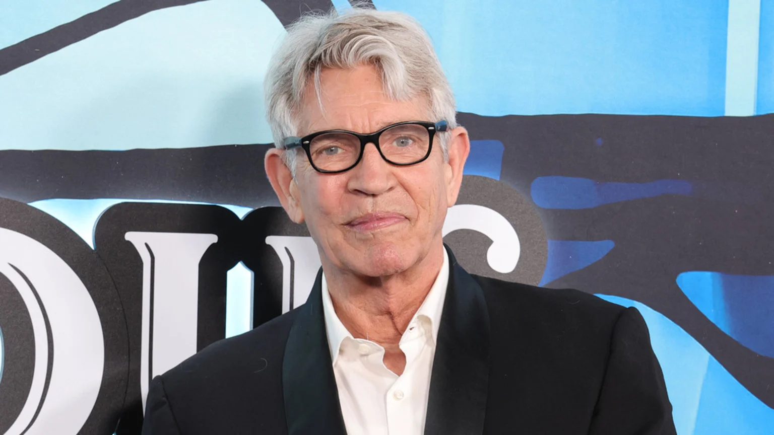Eric Roberts, a man with gray hair and black glasses, smiles gently at the camera while wearing a white shirt and black suit jacket against a blue and black patterned background.
