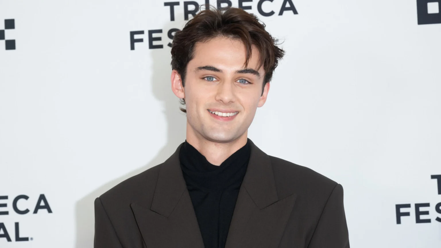 A young man with dark wavy hair and blue eyes smiles at the camera, wearing a dark suit and a black turtleneck against a white background with "TRIBECA FEST" branding.