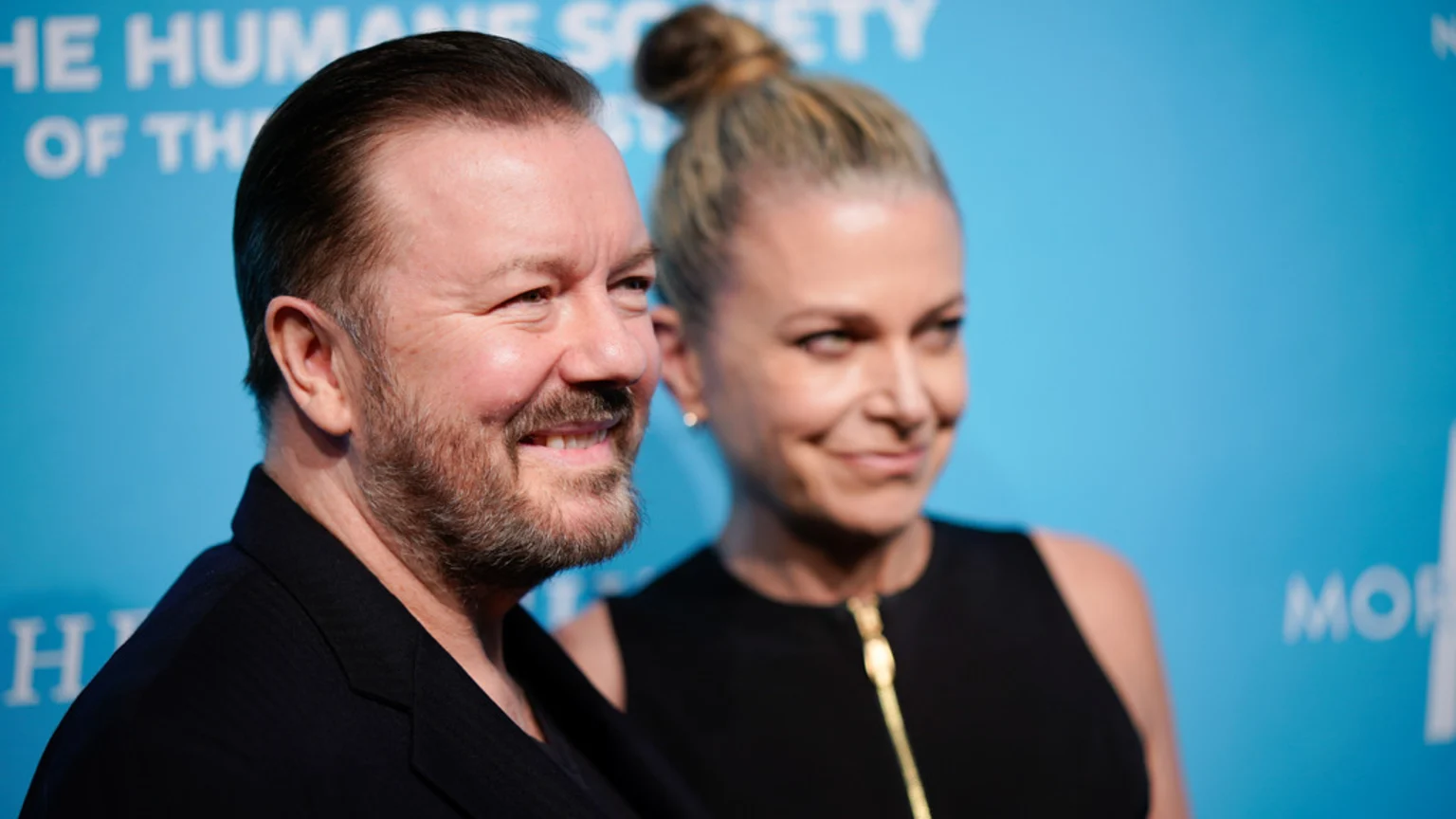 Ricky Gervais smiles in the foreground, wearing a dark jacket, next to his partner Jane Fallon, who is out of focus in a black top with a gold zipper against a blue background.