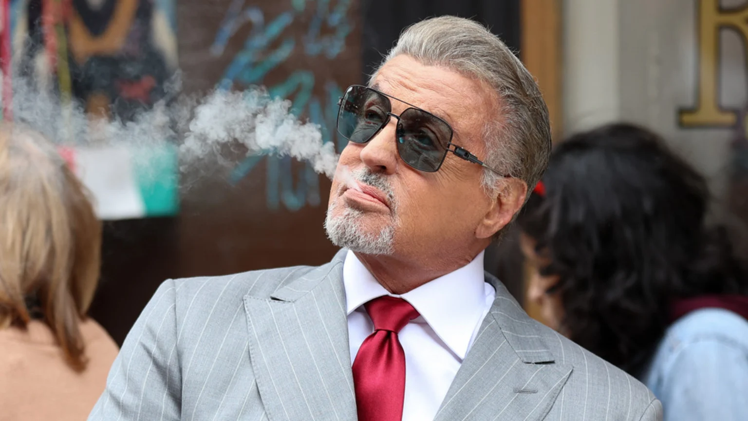 Sylvester Stallone, in a grey pinstripe suit, white shirt, and red tie, exhales smoke while wearing dark sunglasses, looking upwards.