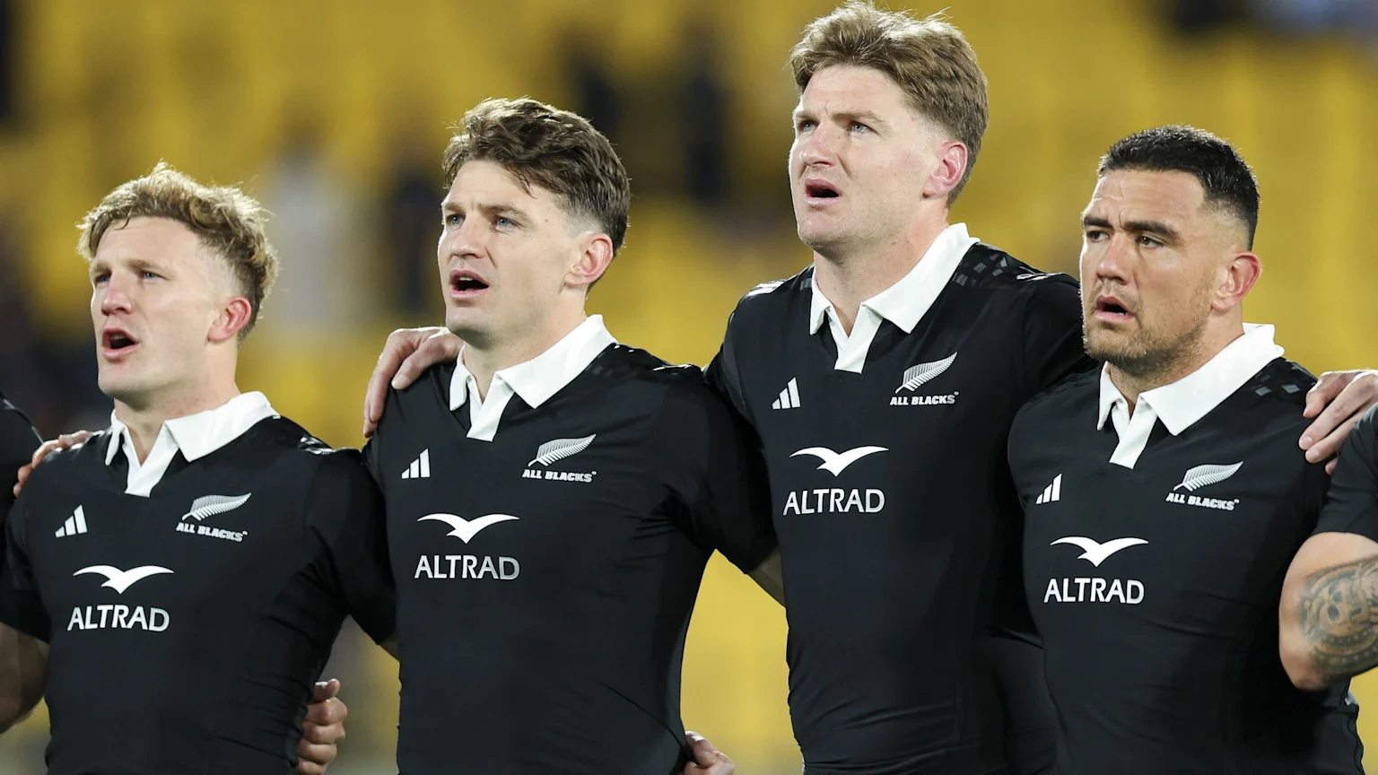 Four All Blacks rugby players in black jerseys with white collars stand side-by-side, appearing to sing an anthem in a stadium.