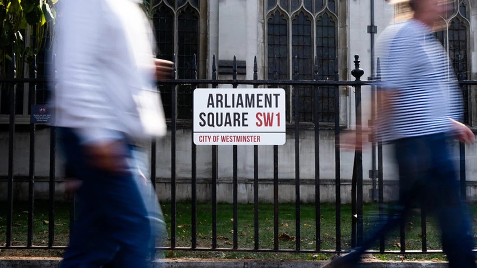 A white street sign on a black fence reads "ARLIAMENT SQUARE SW1 CITY OF WESTMINSTER," with two blurry figures of people walking past on either side and an old building in the background.