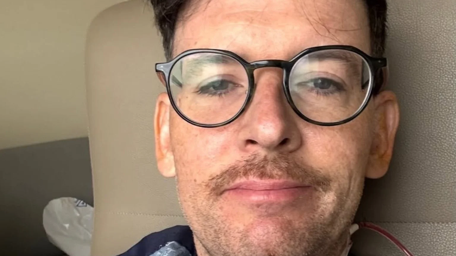Comedian Moshe Kasher, with dark hair, round glasses, and a mustache, looks at the camera with a slight smile; a reddish medical tube is visible near his right shoulder.