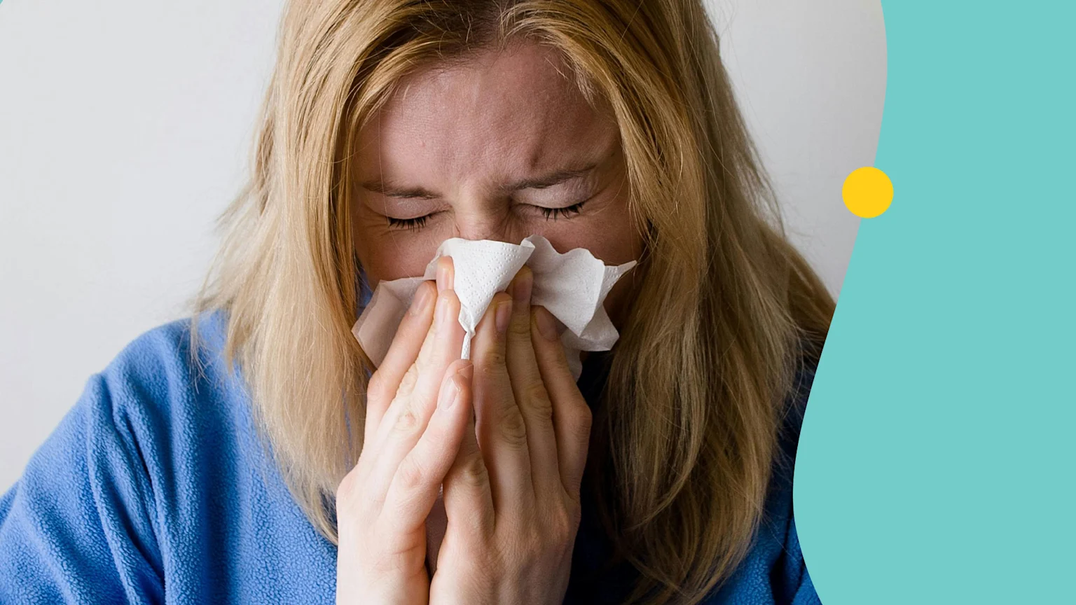 A blonde woman in a blue top holds a white tissue to her nose, with eyes closed and face scrunched, appearing to sneeze or blow her nose.