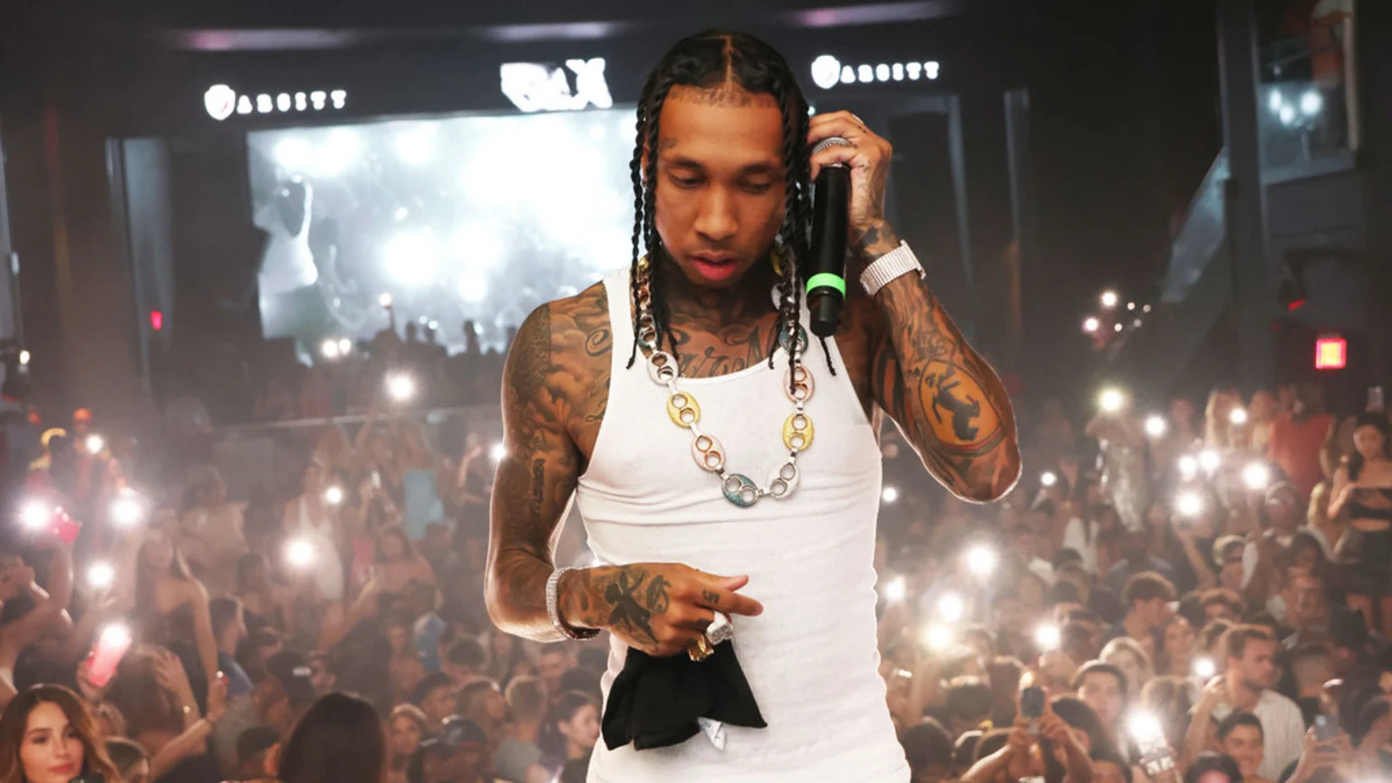 Rapper Tyga performs on stage, holding a microphone, wearing a white tank top and colorful chain, in front of a large crowd illuminating him with cell phone lights.