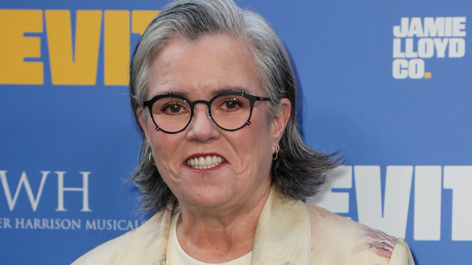 Rosie O'Donnell, a woman with gray hair and black glasses, smiles slightly, wearing a light top and patterned blazer, in front of a blue background with visible text.